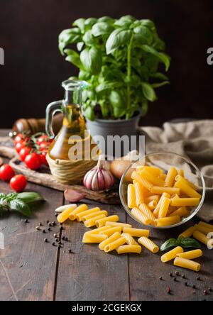 Pâtes de penne crues dans un bol en verre avec de l'huile et de l'ail, des plantes de basilic et des tomates, du poivre et du linge de maison sur fond de table en bois. Banque D'Images