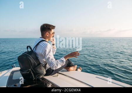 Espace pour le texte. Beau jeune homme avec sac à dos dans les mains, vêtu de vêtements de bureau relaxant et travaillant sur une tablette numérique sur un arc de bateau à moteur, au-dessus de la mer Banque D'Images