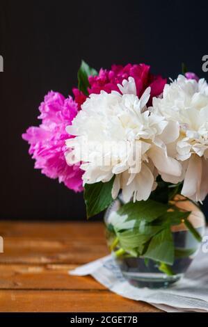 image verticale un bouquet de pivoines fraîches et luxuriantes multicolores dans un élégant vase en verre sur une table en bois contre un mur noir, mise au point sélective Banque D'Images