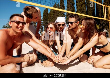 Groupe d'amis souriants et relaxants dans une tenue de plage assis sur le sable formant un cercle, les mains des gens, les caucus empilés les uns sur les autres - les membres de l'équipe, l'amitié Banque D'Images