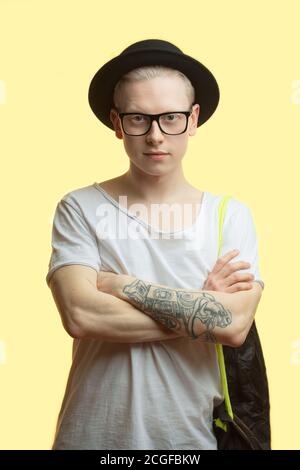 Concept de voyage. Studio portrait de beau jeune photographe homme en chapeau noir, posant avec des bras croisés tatoués. Isolé sur fond jaune. Banque D'Images