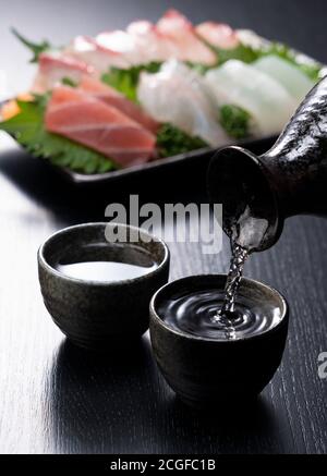 Le saké est versé dans une tasse de saké sur fond noir. Sashimi à l'arrière. Banque D'Images