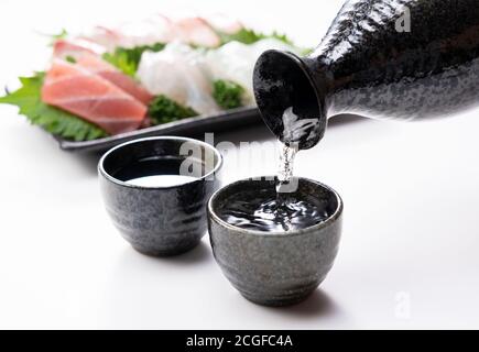 Le saké est versé dans une tasse de saké fixée sur un fond blanc. Sashimi à l'arrière. Banque D'Images