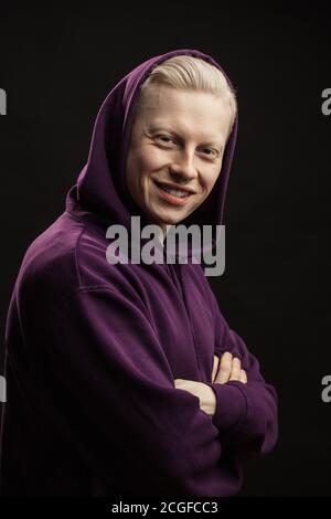 Tête de jeune homme blond heureux en violet sportswear avec capuche sur la tête regardant la caméra, souriant, avec des mains croisées isolées sur le dos noir Banque D'Images