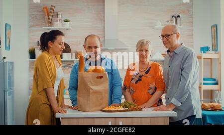 Portrait de famille au ralenti dans la cuisine moderne. Des gens heureux souriant à l'appareil photo dans la salle à manger autour du sac de papier avec des provisions regardant l'appareil photo Banque D'Images