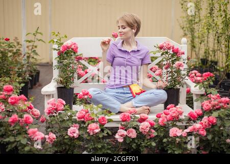 Jeune femme blonde en tenue décontractée d'été assise sur un banc blanc dans un jardin de roses en fleur méditant, appréciant des moments de paix et de calme Banque D'Images