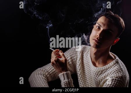 triste homme beau avec la cigarette regardant la caméra dans l'obscurité avec une bouffée de fumée Banque D'Images