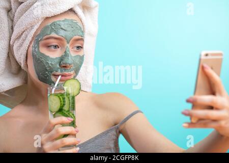 Femme mignonne avec une serviette sur la tête, s'amusant tout en s'arrangeant pour une fête de danse, faisant selfie avec le masque de beauté d'argile sur le visage, plaçant la photo dans soci Banque D'Images