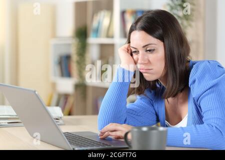 Femme ennuyeuse qui vérifie le contenu en ligne d'un ordinateur portable, assise sur un bureau à la maison Banque D'Images