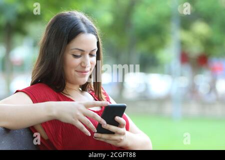 Une femme smiley en rouge vérifie que le smartphone est assis sur un banc dans un parc Banque D'Images