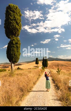 Une femme se tient au milieu d'une route de campagne bordée de cyprès pour prendre une photo. Paysage toscan avec ciel bleu et fond de nuages. Banque D'Images