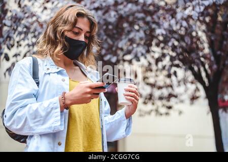 redhead jeune femme portant un masque hygiénique noir debout rue vérifiant son smartphone tout en tenant une tasse de retirez le café Banque D'Images