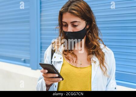 portrait horizontal d'une belle et jeune femme à tête rouge portant un masque noir et l'utilisation d'un smartphone sur une rue avec une ba bleue Banque D'Images