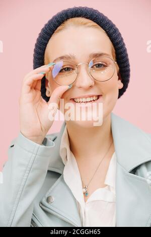 Jeune femme excentrique souriante dans un chapeau et des verres ronds souriant. Sur rose. Banque D'Images