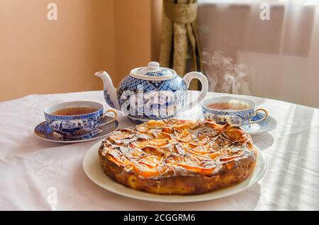 Tarte aux pommes sur un plat blanc, une théière et deux tasses de thé chaud sur une table recouverte d'une nappe blanche. Banque D'Images