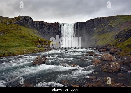 La brume s'élève autour de la base de la chute d'eau de Gufufoss le long de la route menant à Seydisfjordur, Eastfjords, Islande. La rivière se précipite sur des rochers dans son chemin. Banque D'Images