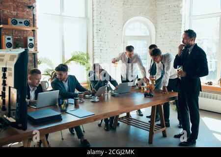 Regarder le football, boire de la bière, encourager. Des collègues heureux et insouciants s'amusent au bureau tandis que leurs collègues travaillent dur et fortement concentrés. Concept de plaisir, de repos, d'occupation professionnelle. Banque D'Images