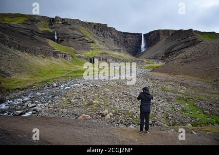 Un randonneur portant des pauses noires pour prendre une photo Le long du sentier de randonnée en direction de la cascade de Hengifoss sur un ciel nuageux Journée grise dans le nord-est de l'Islande Banque D'Images