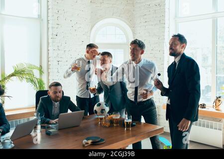 Regarder le football, boire de la bière, encourager. Des collègues heureux et insouciants s'amusent au bureau tandis que leurs collègues travaillent dur et fortement concentrés. Concept de plaisir, de repos, d'occupation professionnelle. Banque D'Images