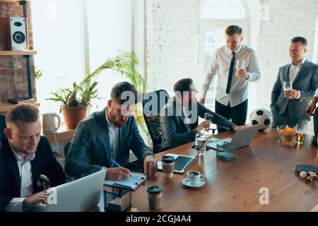 Regarder le football, boire de la bière, encourager. Des collègues heureux et insouciants s'amusent au bureau tandis que leurs collègues travaillent dur et fortement concentrés. Concept de plaisir, de repos, d'occupation professionnelle. Banque D'Images