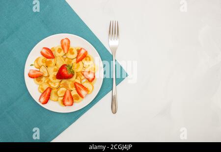 Plats tendance - céréales à crêpes. Tas de mini crêpes aux céréales sur une assiette sur fond blanc avec une serviette turquoise. Fraise et fourchette. Copier l'espace lef Banque D'Images