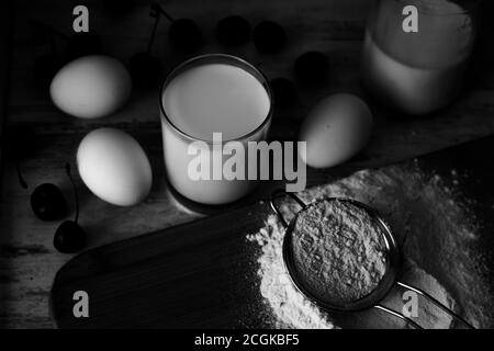 Photo en noir et blanc de la farine dans la passoire métallique, les œufs, le lait et les baies Banque D'Images