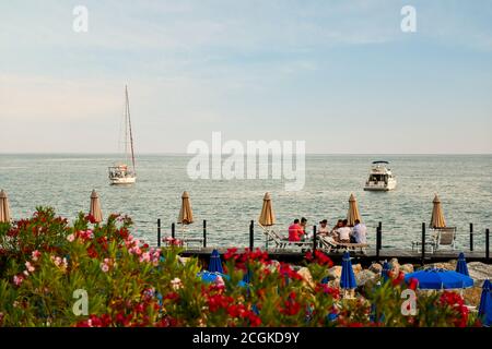 Groupe de jeunes ayant un dîner assis sur des chaises longues sur un quai en bois sur la rive de la mer avec des oléandres fleuries, Lerici, la Spezia, Italie Banque D'Images