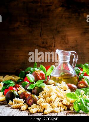 Pâtes italiennes sèches spirales, basilic vert, huile d'olive dans une carafe, tomates cerises noires et rouges, encore la vie dans un style rustique sur fond de bois vintage Banque D'Images