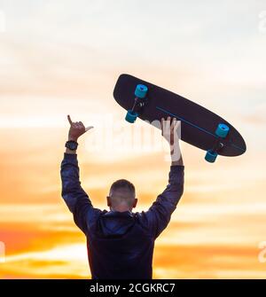 Homme qui profite d'une vie sans soucis au coucher du soleil. Tient le skateboard et montre le panneau Shaka. Banque D'Images