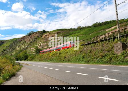 Un train de voyageurs rouge longeant une autoroute vide à côté des collines d'un vignoble en Allemagne. Banque D'Images