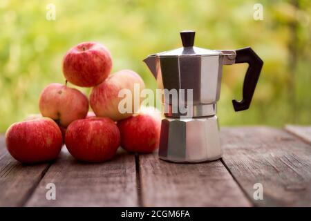 La cafetière geyser repose sur une planche de bois sur un fond vert flou. Avec des pommes rouges Banque D'Images