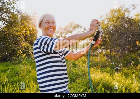 Heureuse femme âgée ayant plaisir arroser des plantes avec le tuyau dans le jardin d'été. Gouttes d'eau dans le rétroéclairage. Agriculture, jardinage, agriculture, vieux Banque D'Images