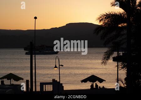 Coucher de soleil sur le golfe d'Aqaba. Des silhouettes d'arbres, des lumières de rue et des personnes sous des parasols de plage sont vues sur la côte jordanienne tandis que deux navires ar Banque D'Images