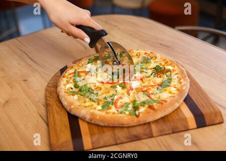 Main femelle avec couteau circulaire coupant des tranches de pizza fraîchement coupées sur une planche de bois dans la cuisine, foyer doux. Trancher à la main la pizza avec un couteau sur la table. Banque D'Images