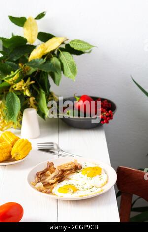 Petit déjeuner copieux sur la table, œufs frits avec du bacon et du maïs bouilli et des légumes, feuilles d'automne dans un vase Banque D'Images