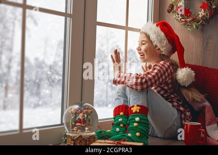 Joyeux Noël et joyeuses fêtes! Jolie petite fille assise près de la fenêtre avec une tasse de boisson chaude et regardant la forêt d'hiver. Chambre décorée. K Banque D'Images