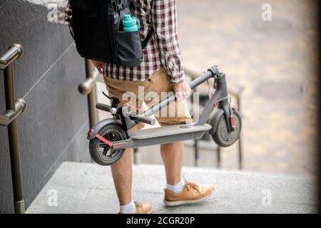 Le jeune homme caucasien en vêtements décontractés avec un sac à dos porte un scooter électrique plié dans sa main jusqu'à l'escalier d'un immeuble de bureau. Homme transportant Banque D'Images