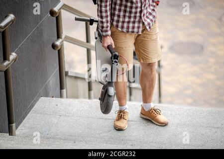 Homme marchant avec un scooter électrique plié en haut des escaliers dans la ville près du bâtiment moderne. Mode de vie technologique écologique. E-mobilité. Personne transportant Banque D'Images