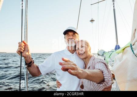 Bonne femme mûre pointant sur la distance tout en s'embrassant son mari sur leur yacht Banque D'Images