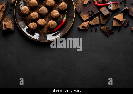 Truffes au chocolat faites maison sur l'assiette avec piments et morceaux de chocolat, fond noir, vue du dessus, espace de copie. Banque D'Images