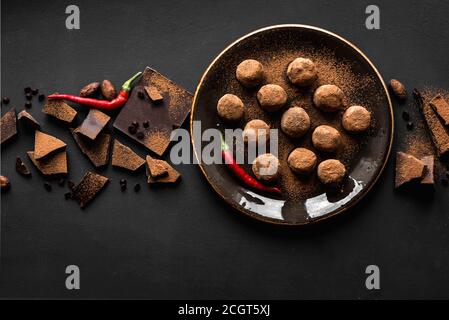 Truffes au chocolat faites maison sur l'assiette avec piments et morceaux de chocolat, fond noir, vue du dessus, espace de copie. Banque D'Images