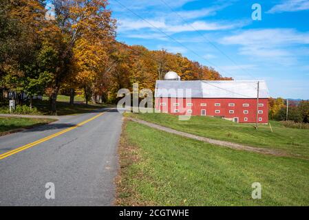 Grange traditionnelle en bois rouge le long d'une route de campagne bordée d'arbres colorés lors d'une journée d'automne ensoleillée. Banque D'Images