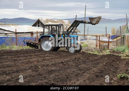Un champ de pommes de terre labouré et un tracteur debout à la clôture contre une maison de village. Banque D'Images