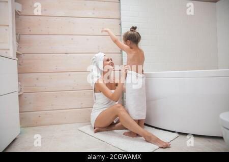Une famille heureuse et aimante qui passe du temps ensemble. Mère et sa fille enfant fille embrassant et embrassant dans la salle de bains blanche. Femme et fille avec des serviettes Banque D'Images