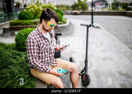 Homme avec e-scooter relaxant et utilisant un smartphone sur banc en ville. Homme moderne avec trottinette assis sur le banc dans le parc tout en discutant sur lui Banque D'Images