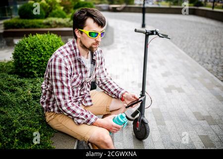 Homme avec e-scooter relaxant et utilisant un smartphone sur banc en ville. Homme moderne avec trottinette assis sur le banc dans le parc tout en discutant sur lui Banque D'Images