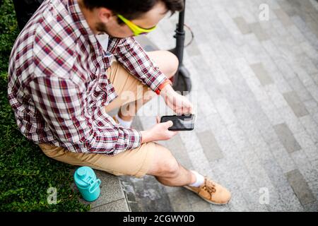 L'homme caucasien utilise les technologies modernes Internet et la communication cellulaire sur smartphone tout en étant assis sur banc dans la ville près du scooter électrique. Écologie Banque D'Images