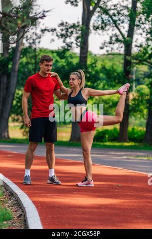 Jeune athlète masculin soutenant sa jeune femme partenaire pendant qu'elle étire les jambes après avoir courir sur une piste de course. Banque D'Images