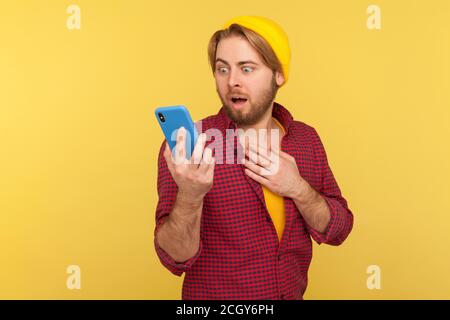 Drôle surpris gars branché dans hipster bonnet beanie et chemise à carreaux soudainement lire le message sur son téléphone mobile et exprimant choc étonnement, unbelie Banque D'Images