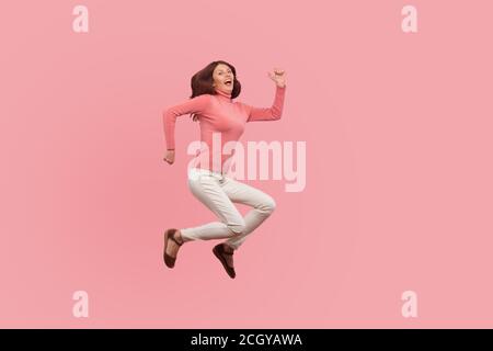 Extrêmement heureuse femme satisfaite avec les cheveux bruns sautant haut ou volant dans l'air, les rêves deviennent réalité. Studio d'intérieur isolé sur fond rose Banque D'Images
